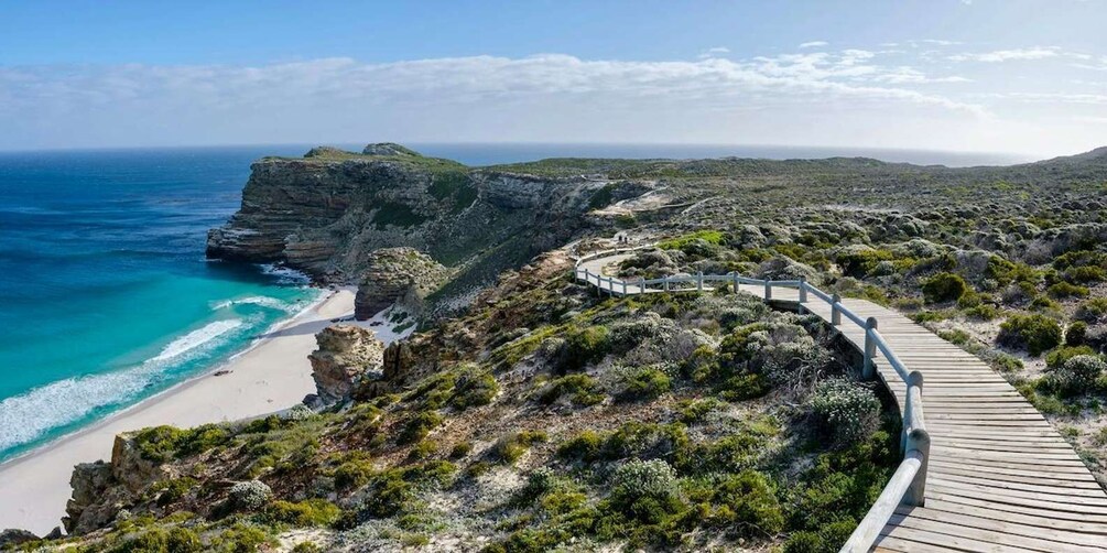 A coastal landscape with a wooden boardwalk leading to a beach, surrounded by rocky cliffs and sparse vegetation. 5