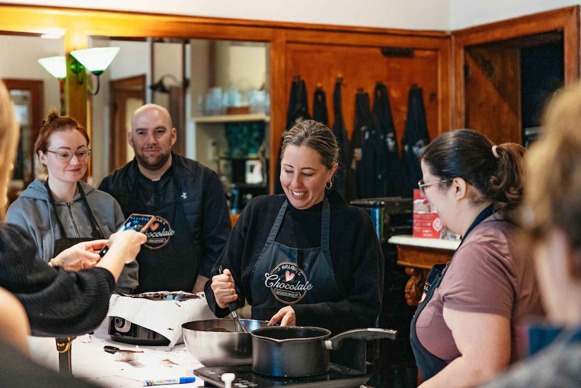 A group of people in a kitchen, some wearing aprons, with a pot on the stove and a box of chocolate on the table. 1