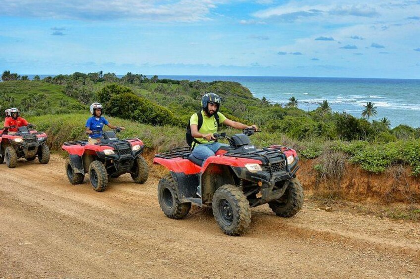 Three people riding red ATVs on a dirt road with a coastal landscape in the background. 3