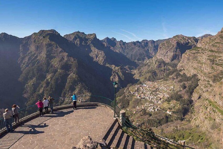 Paesaggio montuoso con una terrazza panoramica pavimentata, persone che ammirano il panorama e un piccolo insediamento nella valle. 1
