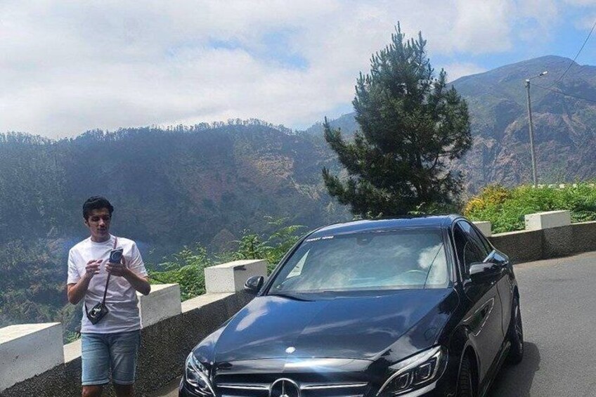 A person standing next to a black car on a road with a mountainous landscape in the background. 4