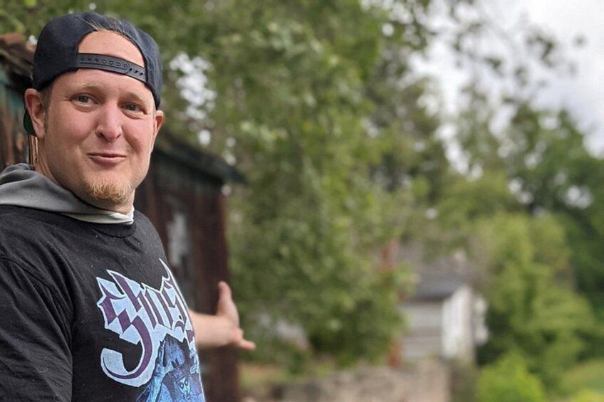 A person wearing a cap and a graphic t-shirt, standing outdoors with trees and a building in the background. 3