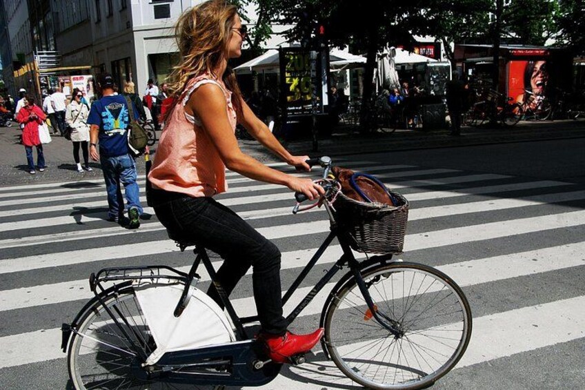 Eine Person fährt mit einem Fahrrad, an dessen Vorderseite ein Korb befestigt ist, über eine Fußgängerampel in einer städtischen Umgebung. 1