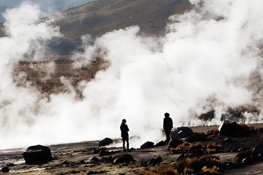 Two people standing on a rocky terrain with steam rising from the ground, surrounded by a barren landscape with distant hills. 1