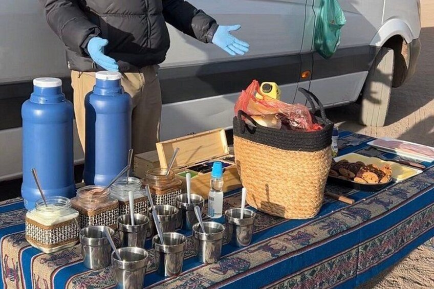 A person standing by a table with food items and containers, a vehicle in the background, and a patterned carpet. 3