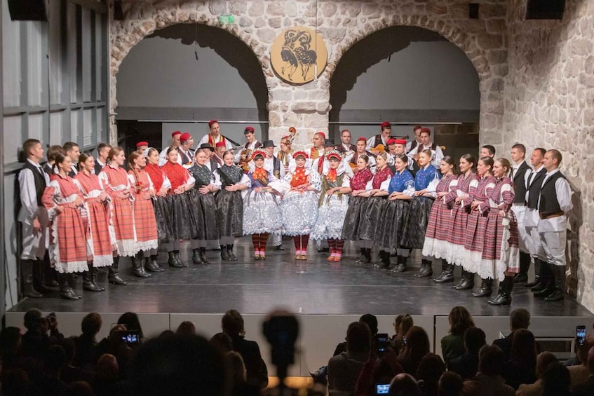A group of people in traditional attire on a stage with stone arches and a coat of arms in the background. 5