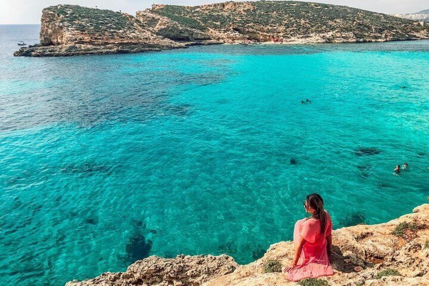 A person in a pink dress sitting on a rocky outcrop overlooking turquoise waters with swimmers in the distance. 4