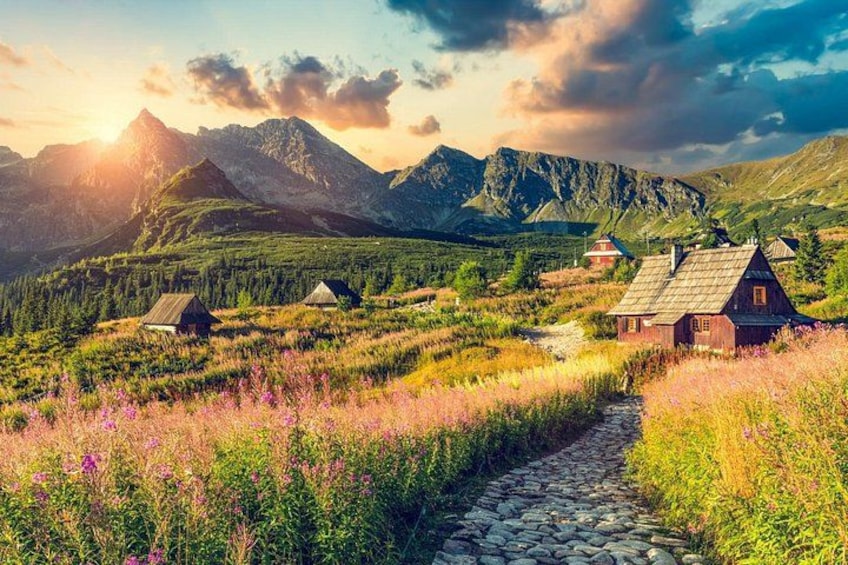 A rural landscape with a stone path, wooden houses, and mountains in the background. 3