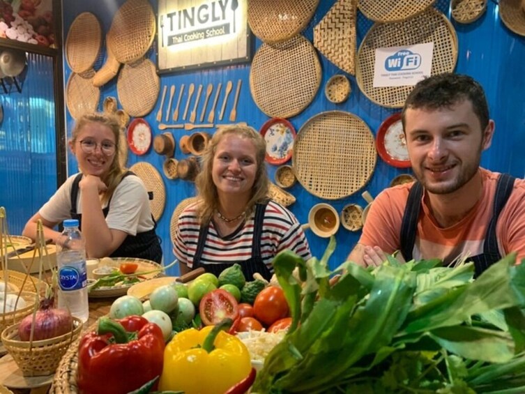 Henkilö istuu pöydän ääressä, pöydällä on erilaisia vihanneksia, ja taustalla on sininen seinä, jossa on näkyvillä kudottuja esineitä. 2