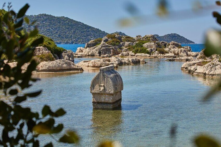 A clear blue sea with a rocky shoreline, a large rock formation in the center, and a hill in the background. 3