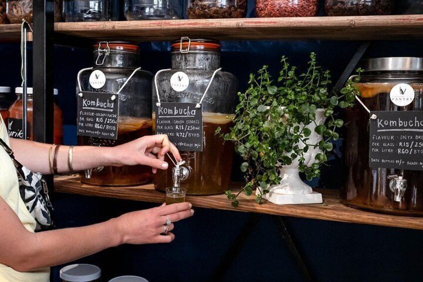 Une personne verse du kombucha d’un bocal dans un verre. On peut voir plusieurs bocaux de kombucha sur une étagère derrière elle. 2