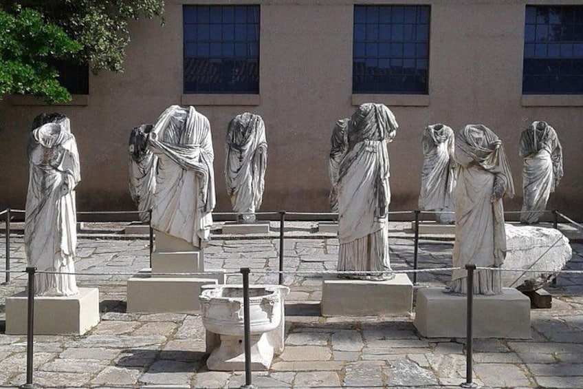 Des statues de marbre représentant des femmes vêtues à la mode classique, disposées dans une cour dallée de pierre, avec un bâtiment en arrière-plan. 4