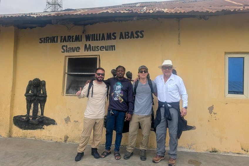 Een groep mensen staat voor een gebouw met de tekst 'Seriki Farea Williams Abass Slave Museum'. 3