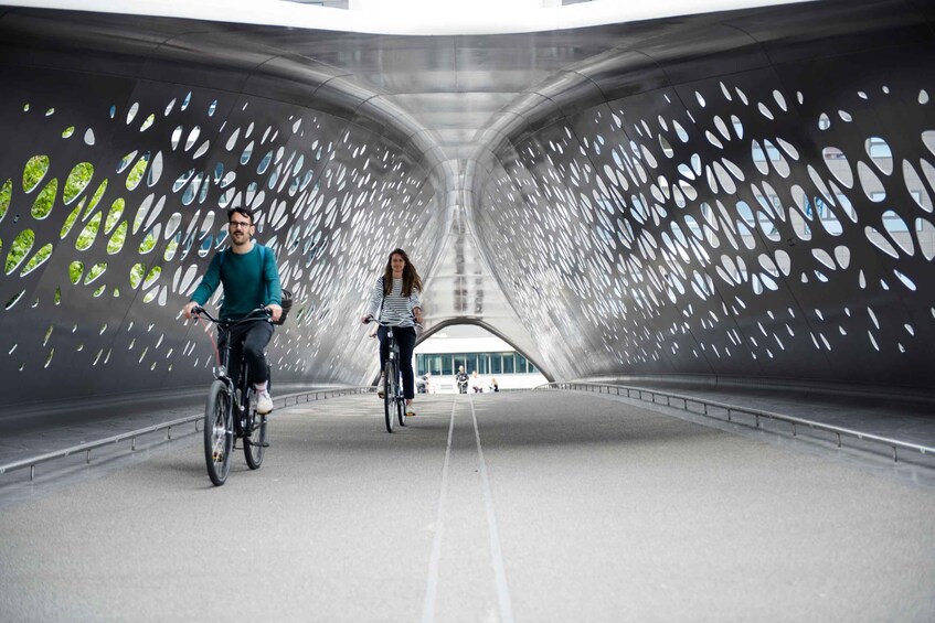 En moderne bro med et unikt perforeret design, to cyklister og en bygning i det fjerne. 1