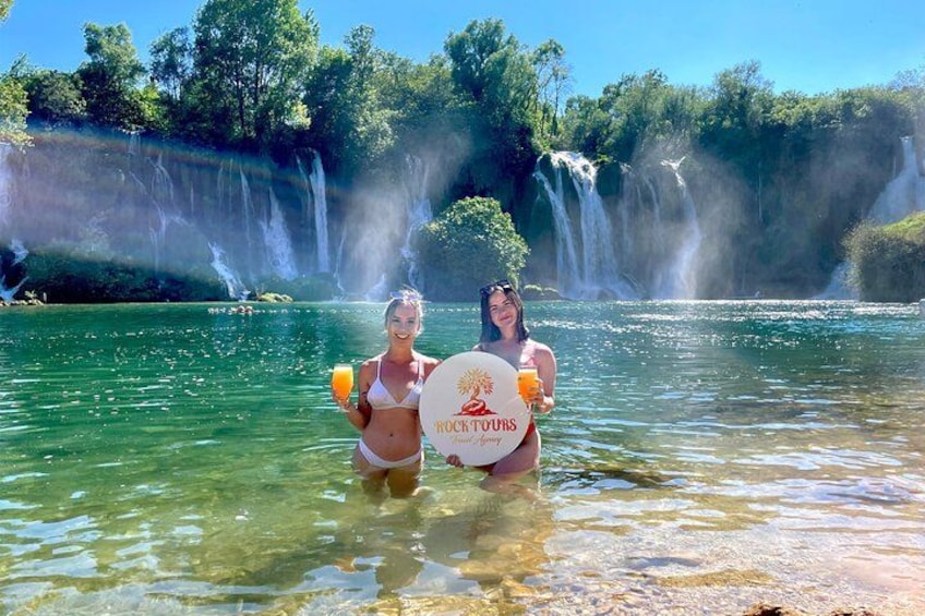 Two people in a clear river with waterfalls in the background. 3