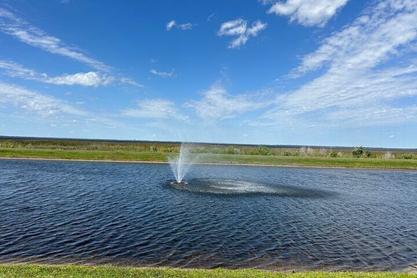 Un plan d’eau paisible, avec une fontaine au centre, bordé de rives verdoyantes et surplombé d’un ciel bleu clair. 4