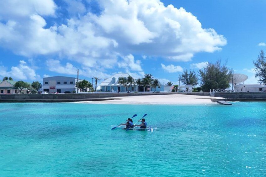 Two people kayaking on a turquoise sea with a beach and buildings in the background. 3