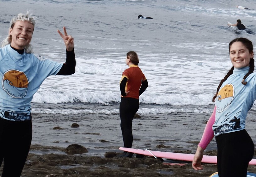 Three people on a beach with surfboards, ocean waves, and other surfers in the background. 2