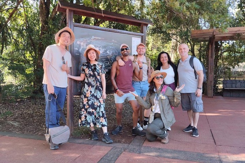 A group of people posing in front of a signpost in a park with trees and a wooden structure in the background. 2