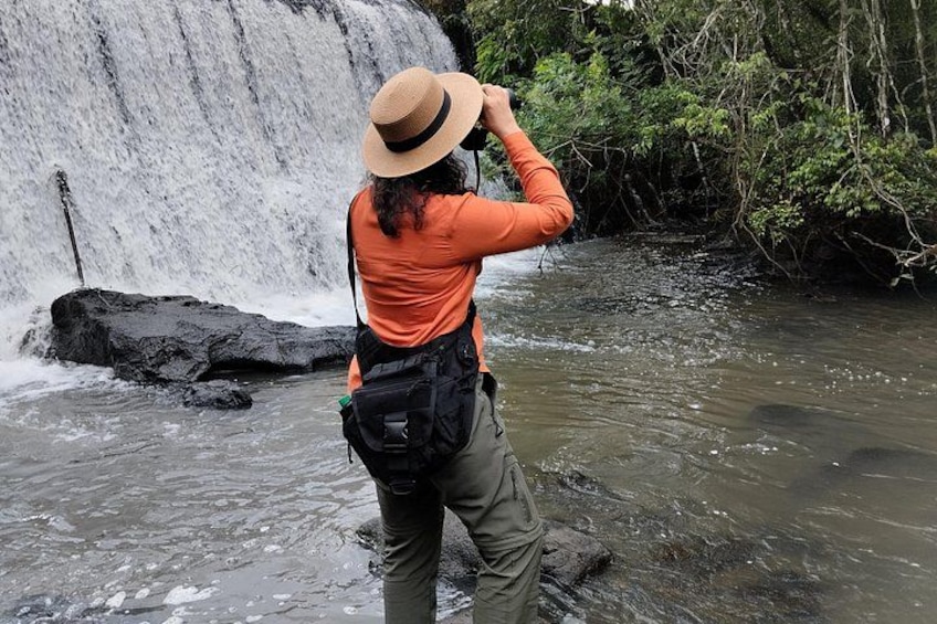 A person in an orange shirt and hat standing in a river with a waterfall in the background. 2