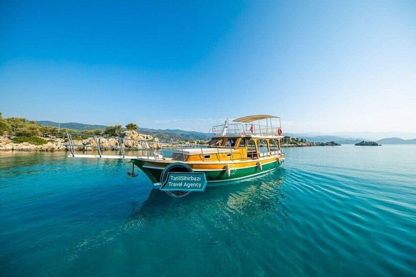 En båd med teksten 'TattiShiraz Travel Agency' på siden, der flyder på roligt blåt vand under en klar himmel med fjerne bakker. 1