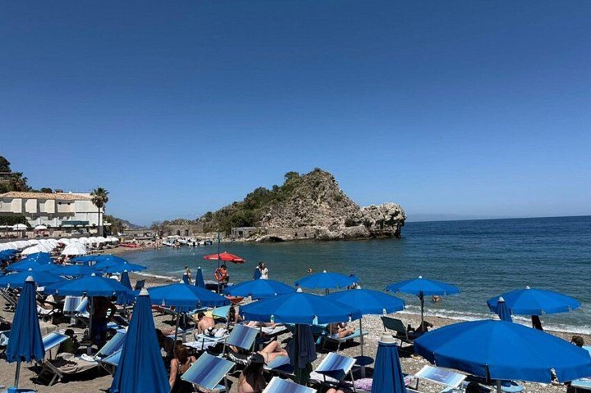 Een strand met blauwe parasols en ligbedden, een rotsachtige richel en een rustige zee. 2