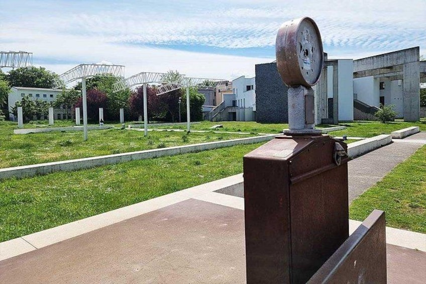 A metal structure with a clock, surrounded by grass and a paved area with buildings in the background. 4