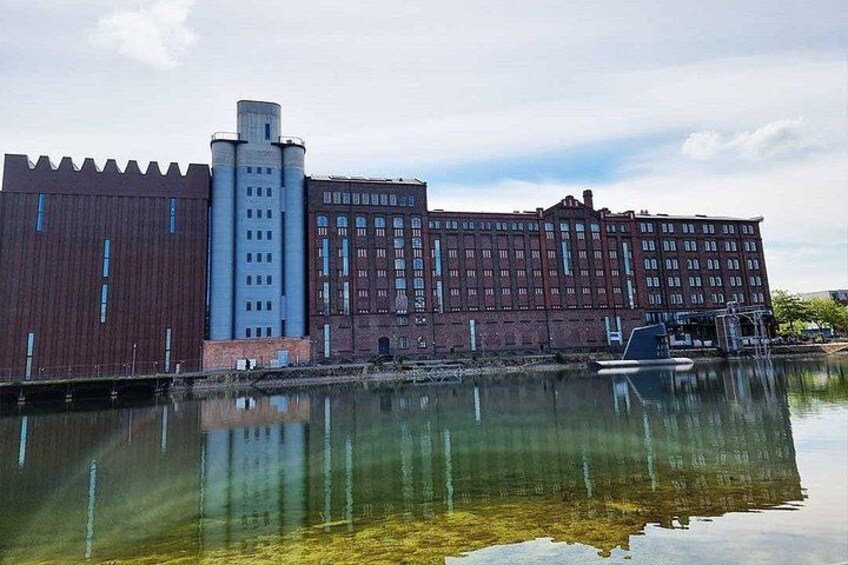 A large industrial building with a tall chimney, a red brick facade, and a body of water reflecting the structure. 2