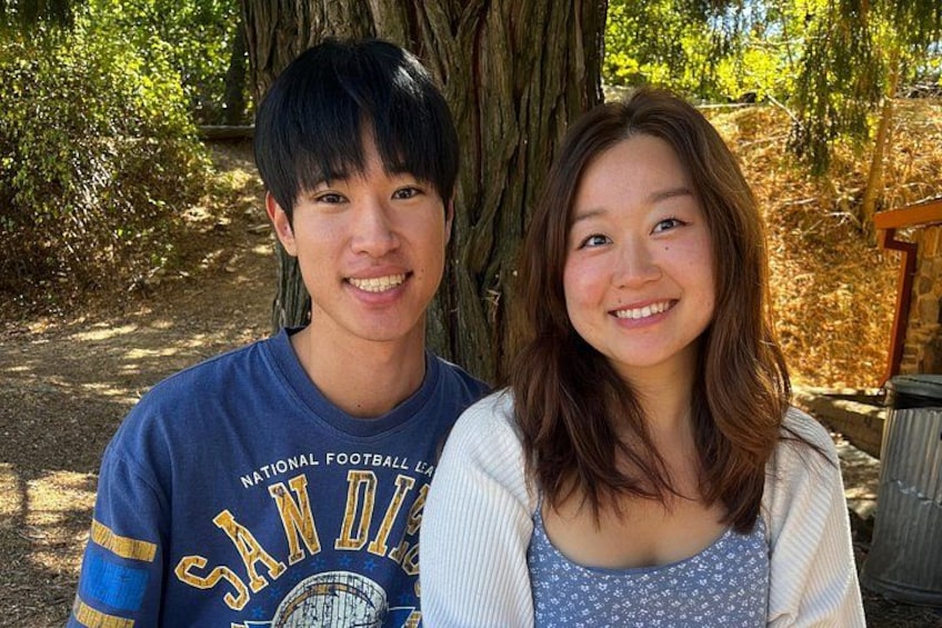 Two people standing in a wooded area with a tree trunk and a wooden structure in the background. 4