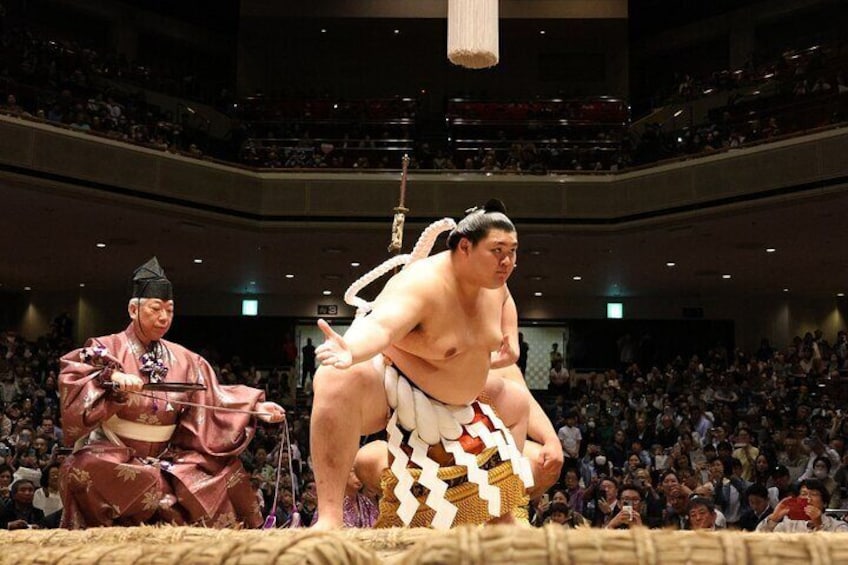 A sumo wrestling match with a referee and a large audience. 5