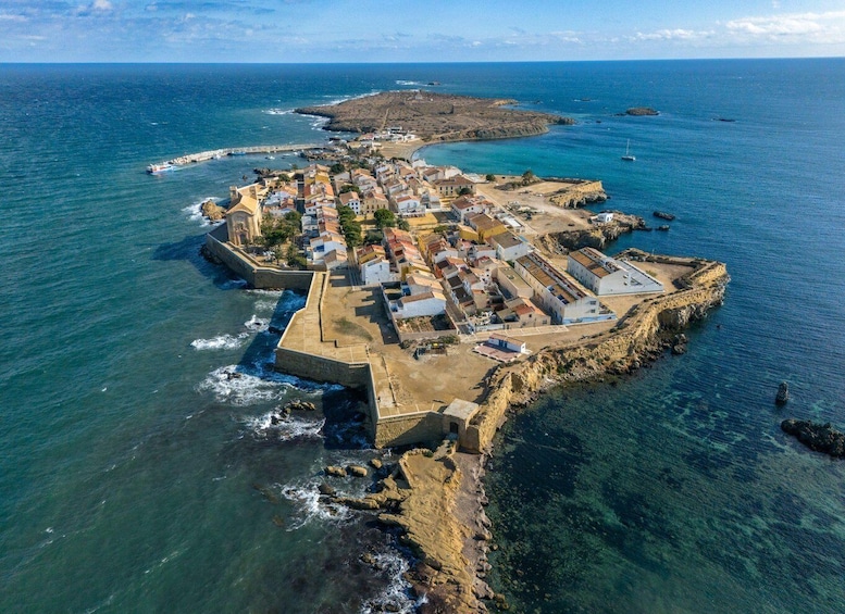 Vista aérea de un pueblo costero con un puerto fortificado, rodeado por el mar y una isla distante. 1
