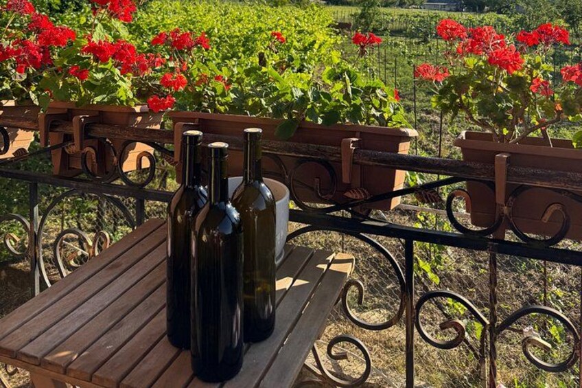 A wooden table with wine bottles and a metal railing with red flowers in the background. 3