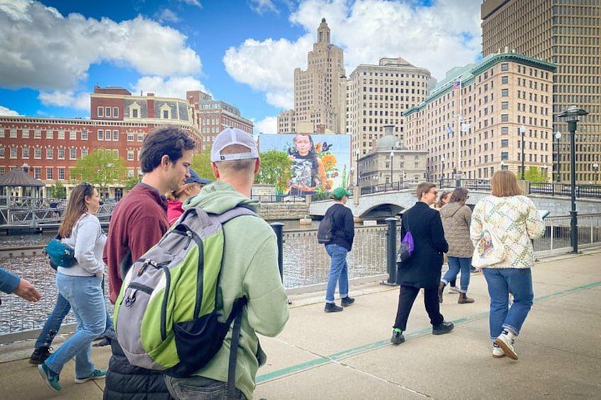 A group of people walking along a riverside walkway with buildings and a mural in the background. 1