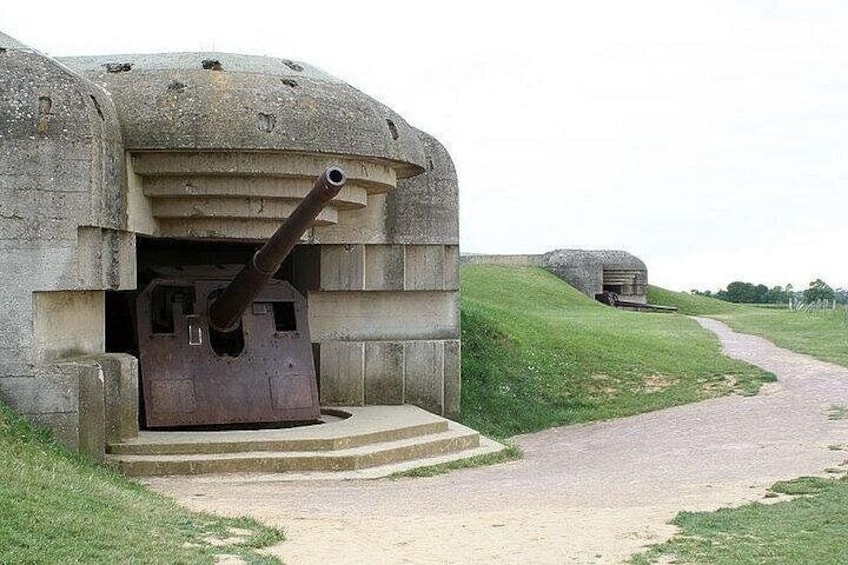 Un bunker in cemento con un grosso cannone sporgente dalla parte anteriore, un sentiero che vi conduce e un altro bunker sullo sfondo. 2