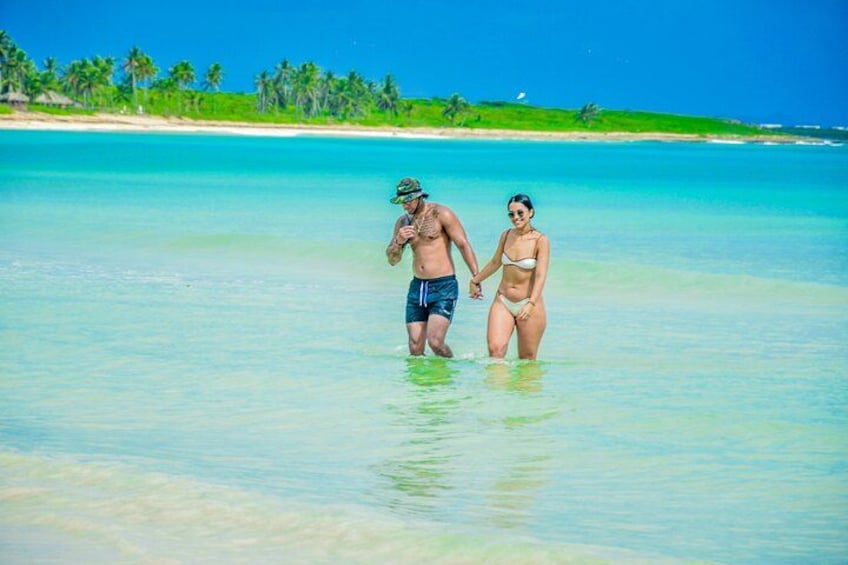A couple walking in shallow clear water with a beach and palm trees in the background. 2