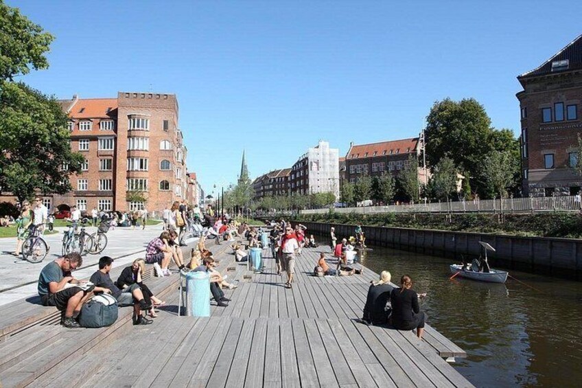 A wooden pier with people sitting and walking, a canal with a boat, and buildings in the background. 5