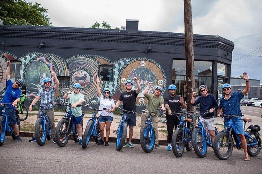 En gruppe mennesker på cykel foran en bygning med et muralmaleri. 1