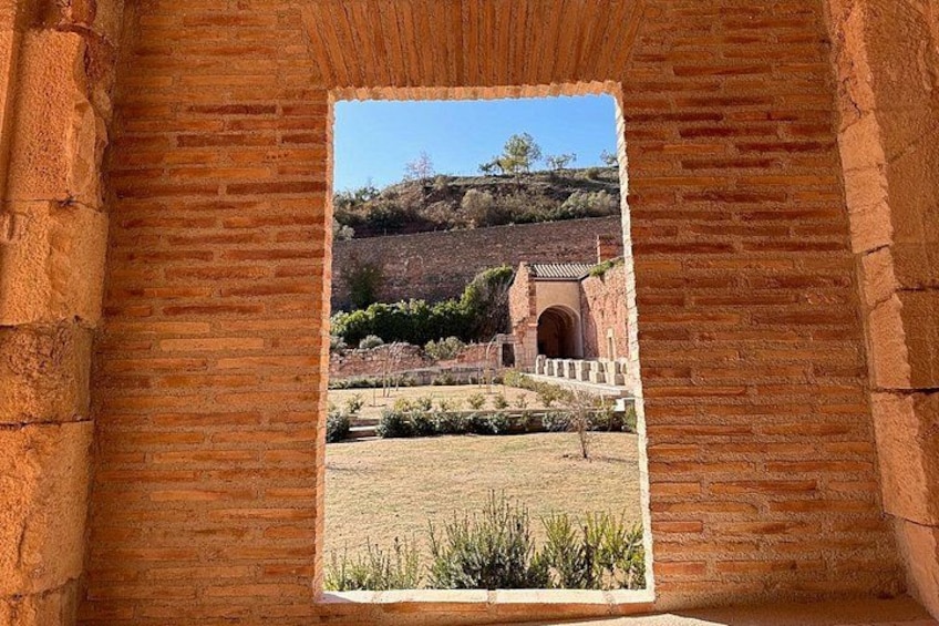 A brick structure with a window overlooking a landscape with a hill, a building, and a pathway. 2