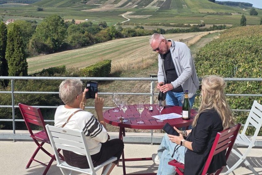 A vineyard with a table and chairs, a person pouring wine, and a person taking a photo. 1