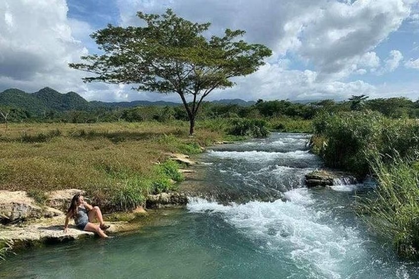 Un río con aguas cristalinas, una persona sentada sobre una roca y un árbol al fondo. 3