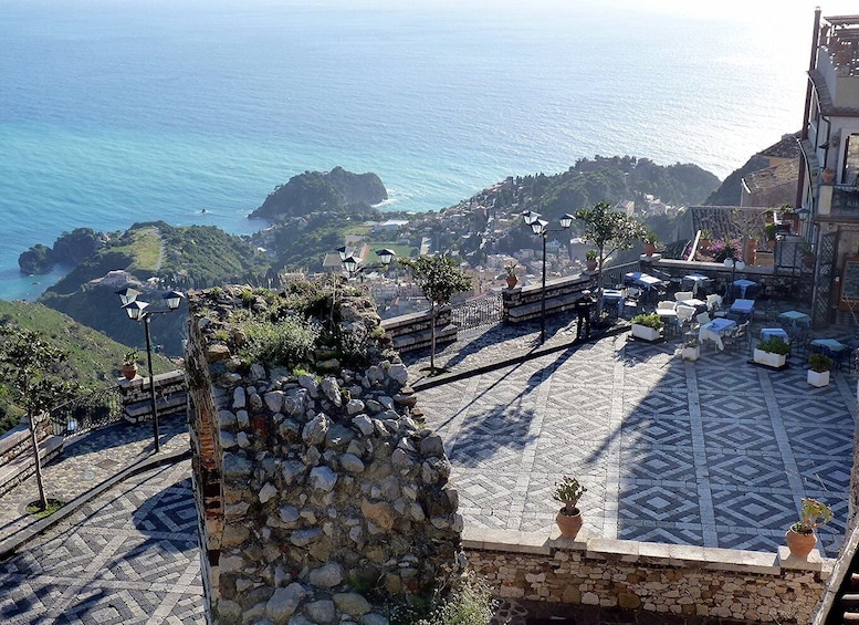 Eine Küstenstadt mit einem gepflasterten Platz, Außenbestuhlung und Blick auf das Meer und die Hügel. 1