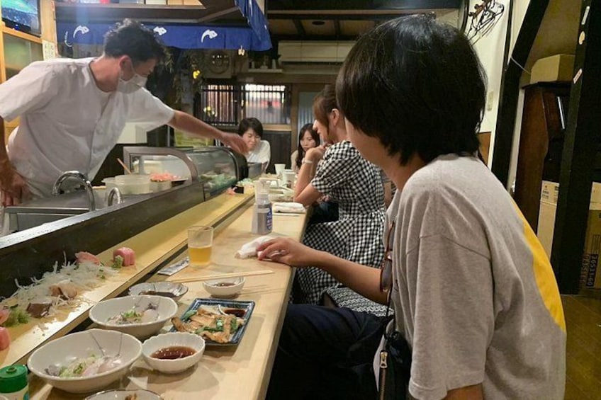 A sushi bar with a chef preparing food, customers seated at tables, and various dishes and condiments on the counter. 4