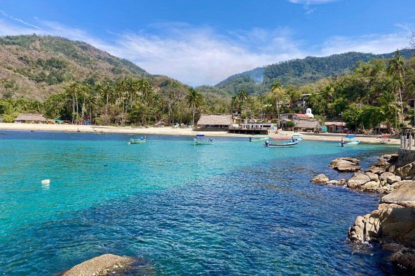 A coastal scene with clear turquoise water, a sandy beach, and a backdrop of hills with vegetation. 2