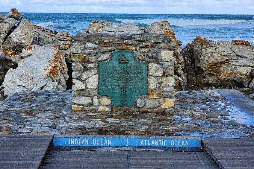 A stone monument with a plaque by the ocean, a wooden walkway, and a rocky shoreline. 1