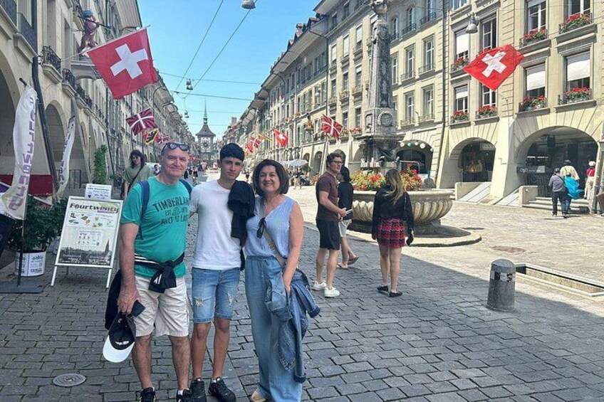 A group of people posing on a cobblestone street with Swiss flags, a fountain, and historic buildings. 3