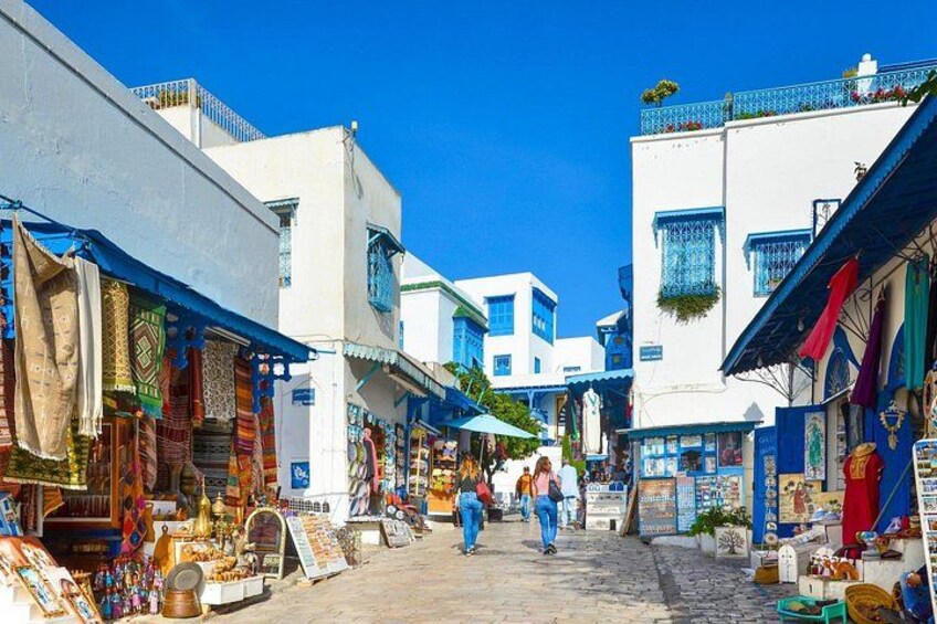 Une rue étroite bordée de boutiques et d’immeubles, où l’on voit des gens se promener, et un ciel bleu dégagé. 4
