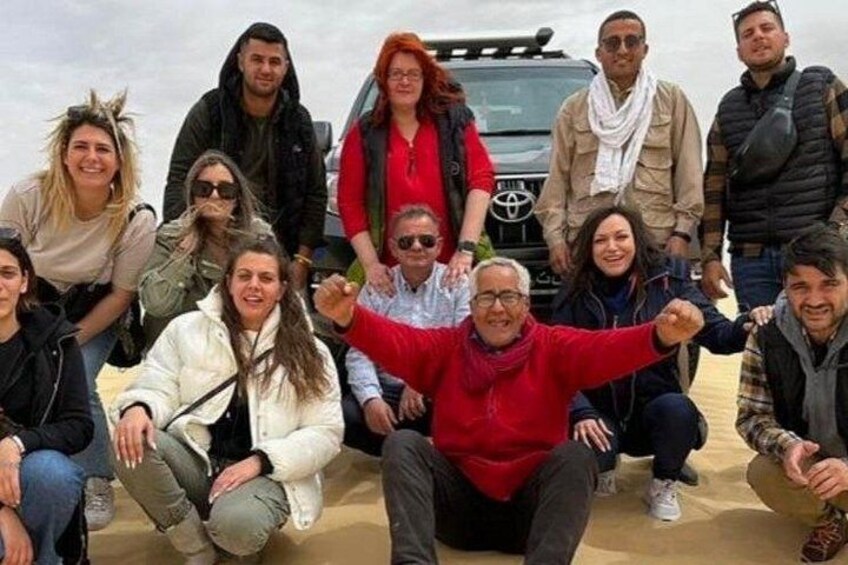 Un groupe de personnes posant sur une surface sablonneuse, avec un véhicule en arrière-plan. 2