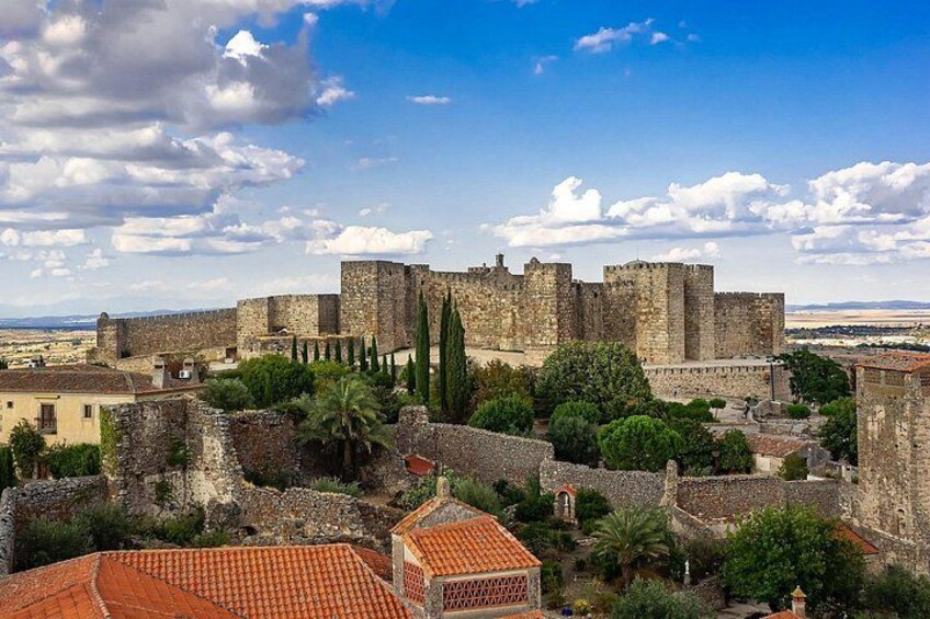 A historic stone castle with a surrounding wall, palm trees, and red-tiled roofs. 2