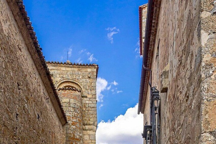 A narrow street with old stone buildings, a clear blue sky, and a prominent tower with an arched entrance. 4
