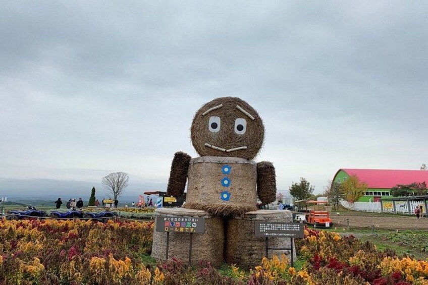 A hay figure with a face and buttons, surrounded by colorful flowers and a sign. 1